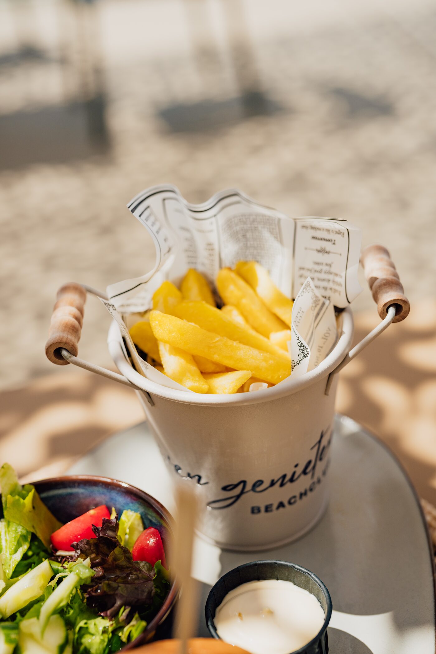 Pommes im Even-Genieten-Beachclub-Eimer mit Salat, im Sand